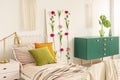 Flower board between single bed with olive green, orange and pastel pink pillows and green wooden cabinet with leaf in glass vase Royalty Free Stock Photo