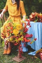 Flowers bouquet of orange, red, yellow dahlias. Brunette woman in yellow dress, maple leaves wreath on wooden chair by round table Royalty Free Stock Photo