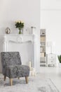 Flowers on fireplace portal in white apartment interior with grey patterned armchair on carpet. Real photo