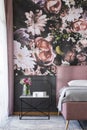 Flowers on table next to pink bed in bedroom interior with patterned wallpaper. Real photo