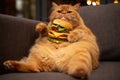 A fluffy ginger cat sits on a sofa and eats a large cheeseburger with lettuce and tomato Royalty Free Stock Photo