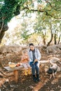 Fluffy piglet stands by a bench with sitting mother and little girl reading a book in the garden Royalty Free Stock Photo