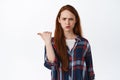 Flustered young redhead girl pointing left and frowning upset, stare confused after showing advertisement, strange promo