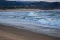 Foaming ocean waves breaking on sandy shore in evening dusk. Surf on beach with village and hills on background. Royalty Free Stock Photo