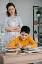 Focus of attentive mother standing with crossed arm behind son sitting at desk and doing homework