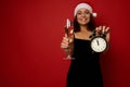 Focus on champagne flute with sparkling wine and alarm clock in the hands of blurred pretty woman in Santa hat, smiling with