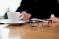 Focus on eyeglasses on table. Blurred concentrated man writing something in his planner. Cup of yummy flat white. Wooden Royalty Free Stock Photo