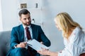 Focus of handsome recruiter holding clipboard and pen while sitting in armchair near blonde employee