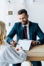 Focus of happy recruiter holding pen near clipboard and looking at blonde employee