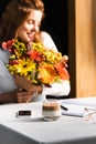 Focus of happy woman with bouquet of autumn flowers in cafe with coffee cup, smartphone and notebook Royalty Free Stock Photo