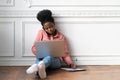 Focused Afro-American woman making notes, talking on phone, using laptop and sitting on floor. Female employee consulting by Royalty Free Stock Photo