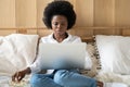 Focused Afro-American woman relaxing after work, eating popcorn while watching horror or thriller movie on laptop, lying in bed.