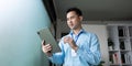 Focused Asian businessman using a tablet for planning, standing by a window in a modern office environment. Royalty Free Stock Photo