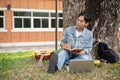 A focused Asian male student is reading a book or writing some ideas in a book under the tree Royalty Free Stock Photo