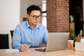 Focused asian middle aged man using laptop and taking notes, writing business report, sitting at table at home Royalty Free Stock Photo