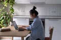 Focused Asian woman freelancer working remotely sits at table with laptop in kitchen Royalty Free Stock Photo