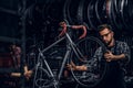 Focused attractive man in glasses is chainging wheel for bicycle at busy workshop