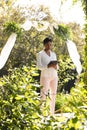 Focused biracial female marriage officiant using tablet wedding arch with flowers in sunny garden Royalty Free Stock Photo