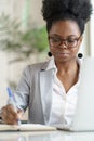 Focused Black businesswoman or employee in glasses, working at laptop at home office, makes notes.