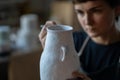 Focused brunette woman paints text with brush on white ceramic vase at table on blurred background