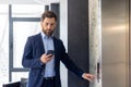 Focused businessman in suit using smartphone while waiting for elevator in modern office building Royalty Free Stock Photo