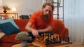 Focused bearded redhead young man playing chess leisure board game alone, domestic activity at home