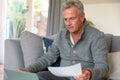 Focused caucasian senior man sitting on sofa, doing paperwork, using laptop