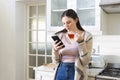 Focused caucasian woman standing, holding tea cup, using smartphone in kitchen at home, copy space Royalty Free Stock Photo