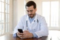 Focused doctor wearing glasses using smartphone, sitting at work desk