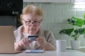 Focused elderly woman in glasses shopping online with credit card using laptop computer at home Royalty Free Stock Photo