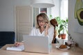 Focused employee woman isitting with dog at home, taking on mobile phone, remote working at laptop