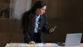 Focused female arabian employee designer business woman standing at office desk using mobile phone checking information