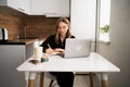 focused female student studies at home, sitting in kitchen with laptop and writing notes in notebook. online learning, self-study Royalty Free Stock Photo