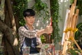 A focused and handsome young Asian man is measuring his drawing on a canvas with a paintbrush