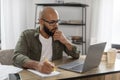 Focused latin man looking at laptop screen and writing in notebook, sitting at workplace at home office, free space