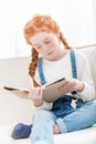 Focused little girl reading book while sitting on sofa at home Royalty Free Stock Photo