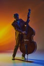 Focused male African-American artist in coat, performing on upright bass, in warm neon light against gradient background Royalty Free Stock Photo