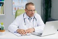 Focused middle aged senior head doctor in white medical coat and glasses sitting at workplace, talking to patient making