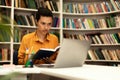 Focused mixed race lady reading book and looking at laptop, studying online, sitting in library, free space Royalty Free Stock Photo