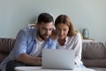 Focused positive millennial married couple sitting on couch at laptop