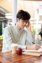 A focused Asian man is jotting down his ideas in a book while sitting at an outdoor table in a cafe Royalty Free Stock Photo