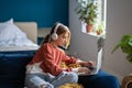 Focused teen girl wearing wireless headphones sitting on sofa with laptop