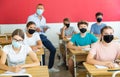 Focused teenage students in protective face masks studying in classroom with teacher, writing lectures in workbooks.