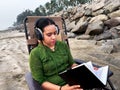 Focused woman with headphones reading book while sitting on a deck chair at a sandy beach location Royalty Free Stock Photo