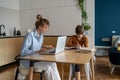 Focused work-at-home mom typing on laptop while sitting at kitchen table with child doing homework Royalty Free Stock Photo