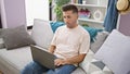 Focused young hispanic man, engrossed in his laptop while working online, sitting, on a cozy sofa at home in a well-illuminated Royalty Free Stock Photo