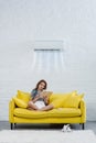 focused young woman reading book on couch under air conditioner hanging on wall and blowing Royalty Free Stock Photo