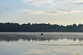 Foggy dawn on the river, a lone fisherman from a boat fishing, forest and clouds reflected