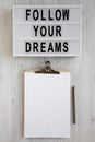 `Follow your dreams` words on a lightbox, clipboard with blank sheet of paper on a white wooden background, top view. Overhead,