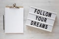 `Follow your dreams` words on a lightbox, clipboard with blank sheet of paper on a white wooden surface, top view. Overhead, fro